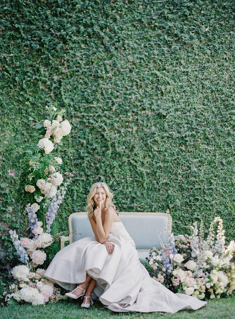 An entire wall of Ivy is the backdrop for this bridal portrait of a bride in a strapless ivory gown with full skirt and train designed by Anne Barge. She is sitting on a vintage pale blue couch with her legs crossed and leaning forward on with her elbow on her knee and hand under chin and laughing. She is surrounded by flower arrangements, one side towering over her, and more on the other side that are as tall as the couch. Photographed at a Charleston wedding by wedding photographers in Charleston Amy Mulder Photography.
