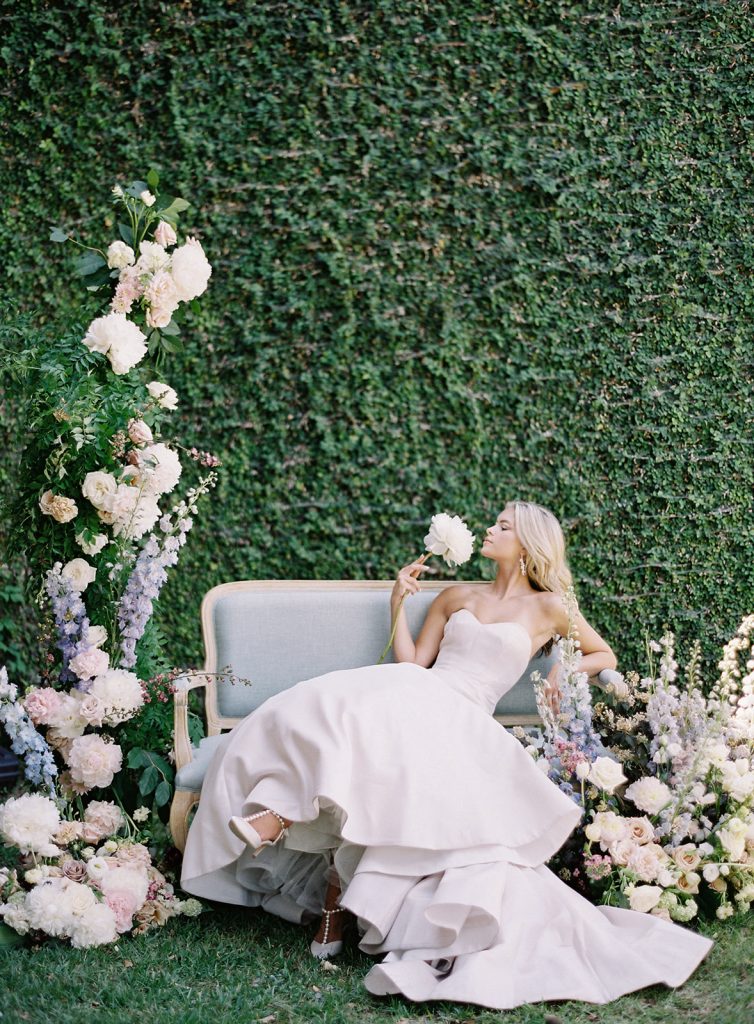 An entire wall of Ivy is the backdrop for this bridal portrait of a bride in a strapless ivory gown with full skirt and train designed by Anne Barge. She is sitting on a vintage pale blue couch with her legs crossed and is surrounded by flower arrangements, one side towering over her, and more on the other side that are as tall as the couch. She is leaning back on her elbow and profile is shown as she has a large flower blossom she is holding up and smelling. Photographed at a Charleston wedding by wedding photographers in Charleston Amy Mulder Photography.