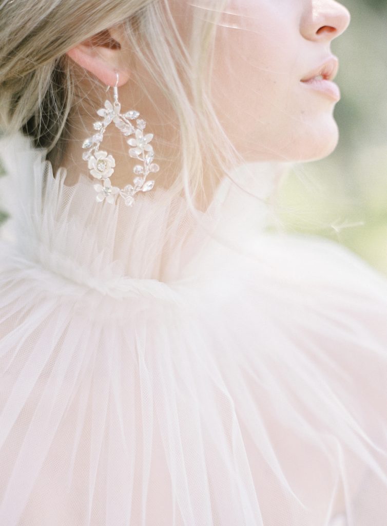 Drop earrings by Eden Luxe Bridal. Tear drop shaped earrings with crystals as leaves and a lite pink flower. Hair is in a low chignon bun with lose locks of hair falling down around her jaw line. With unique sheer collar draping down over her shoulders.  Photographed by wedding photographers in Charleston Amy Mulder Photography