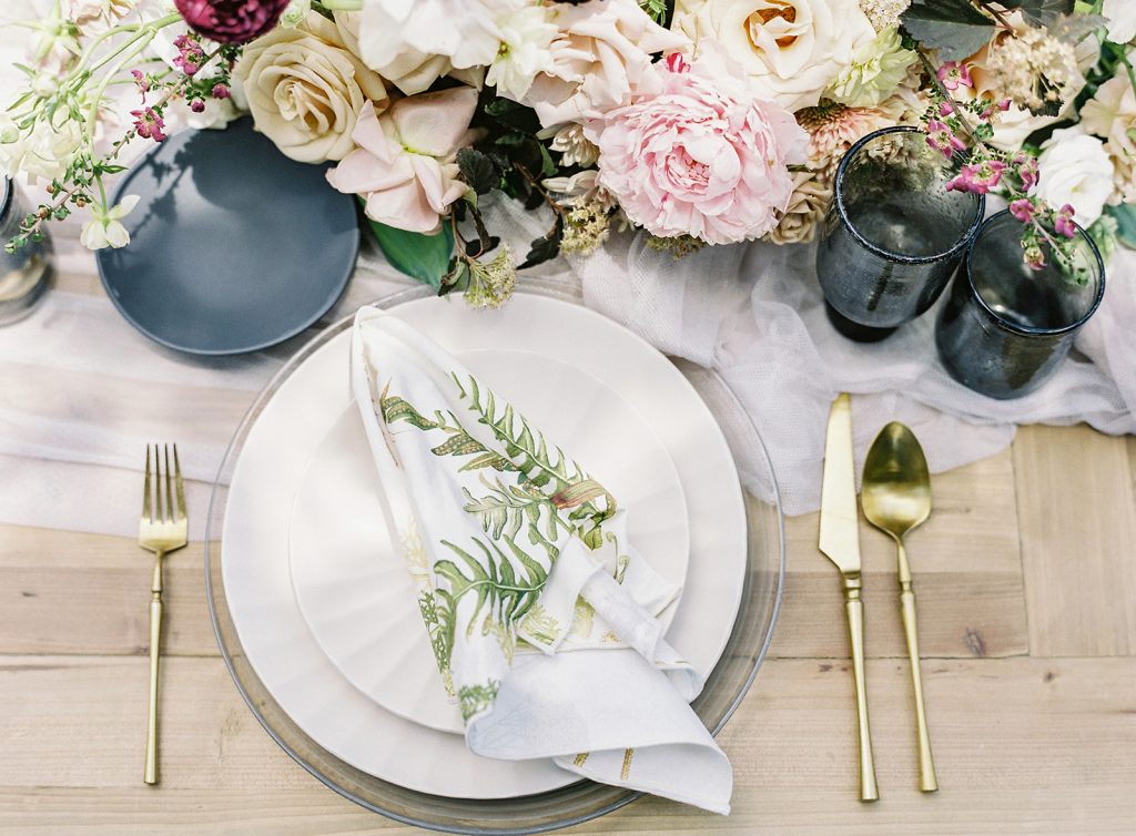 Reception table setting. Ivory plates, gold flatware, black glassware, and pink, taupe, purple and white florals. White napkins with leaves on them. Photographed by wedding photographers in Charleston  Amy Mulder Photography