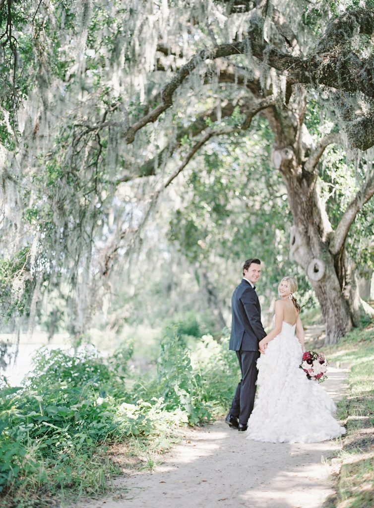 Couple walking down a long narrow path under large oak trees with spanish moss cascading down all around them. They are walking away from the camera, but looking back and smiling. Photographed by wedding photographers in Charleston Amy Mulder Photography.