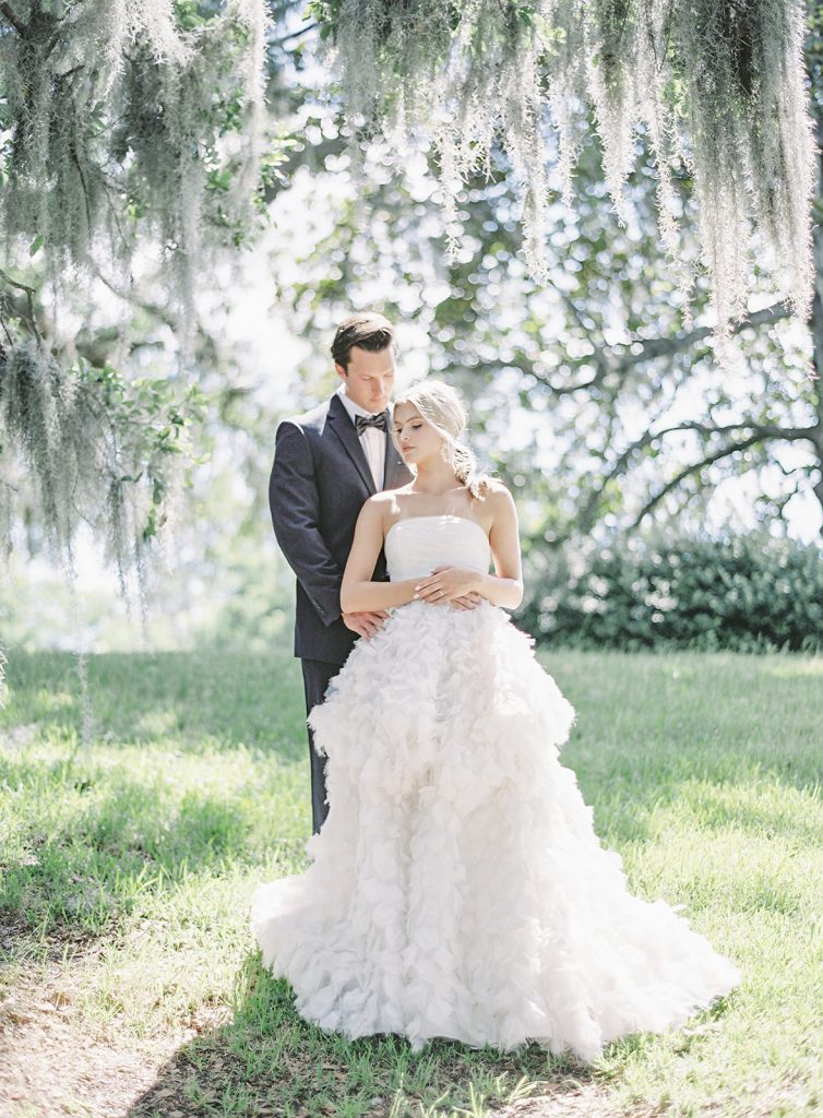 Bride and groom standing and leaning in to each other under an oak trees. Groom is in black tux with black bowtie. Bride is in a strapless ivory wedding gown with fitted bodice and straight neckline and feathered layers of skirt designed by Anne Barge. Photographed by wedding photographers in Charleston Amy Mulder Photography