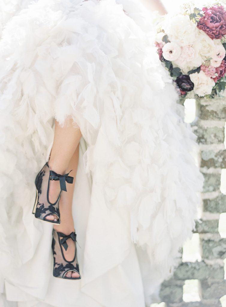 Bride with white feathered gown by Anne Barge and black lace with satin tie wedding shoes by Bella Belle. Bride is sitting on a stone gate with her legs crossed. Photographed by wedding photographers in Charleston Amy Mulder Photography.