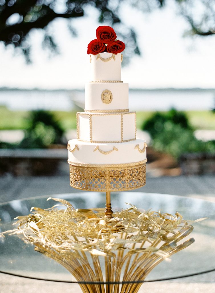 White four tiered wedding cake with gold detailing on top of a gold pedestal surrounded by gold painted leaves on a glass table top with gold table legs. Red garden roses on top of the cake. Photographed at Lowndes Grove by wedding photographers in Charleston Amy Mulder Photography.