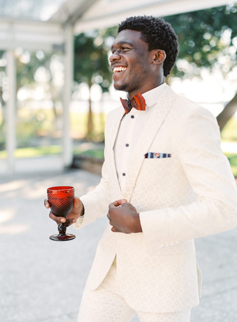 Groom in an ivory and gold detailed tuxedo with red bowtie and holding a red goblet looking out at guests and smiling really big. Photographed at Lowndes Grove by wedding photographers in Charleston Amy Mulder Photography