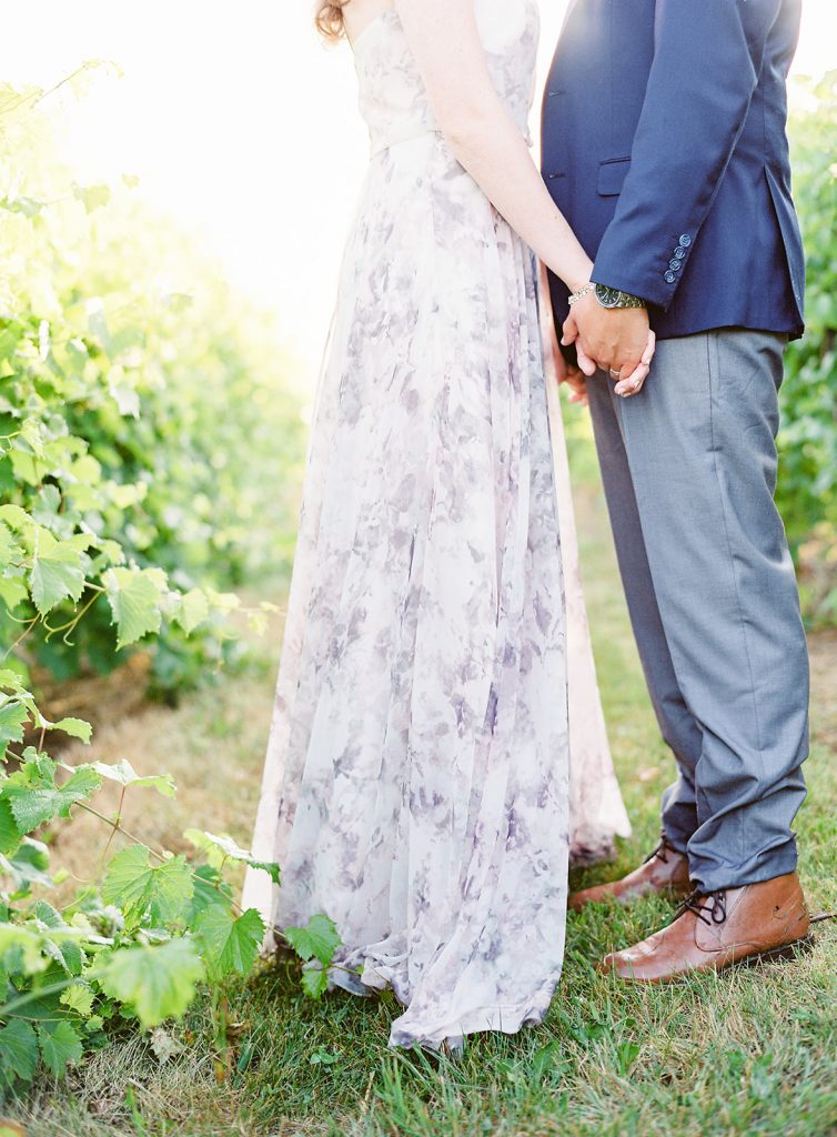 Couple standing facing each other holding each others hands down to their sides. He is in grey suit pants and navy suit jacket, she is in a pastel floral printed floor length dress standing in a row of grape vines at a vineyard. Photographed by wedding photographers in Charleston Amy Mulder Photography.