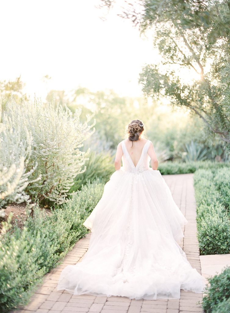 Bride walking away from the camera on a brick path surrounded by cactus and trees with the sun coming in directly behind the trees to give an etherial glow. Photographed at El Chorro Wedding by wedding photographers in Charleston Amy Mulder Photography.