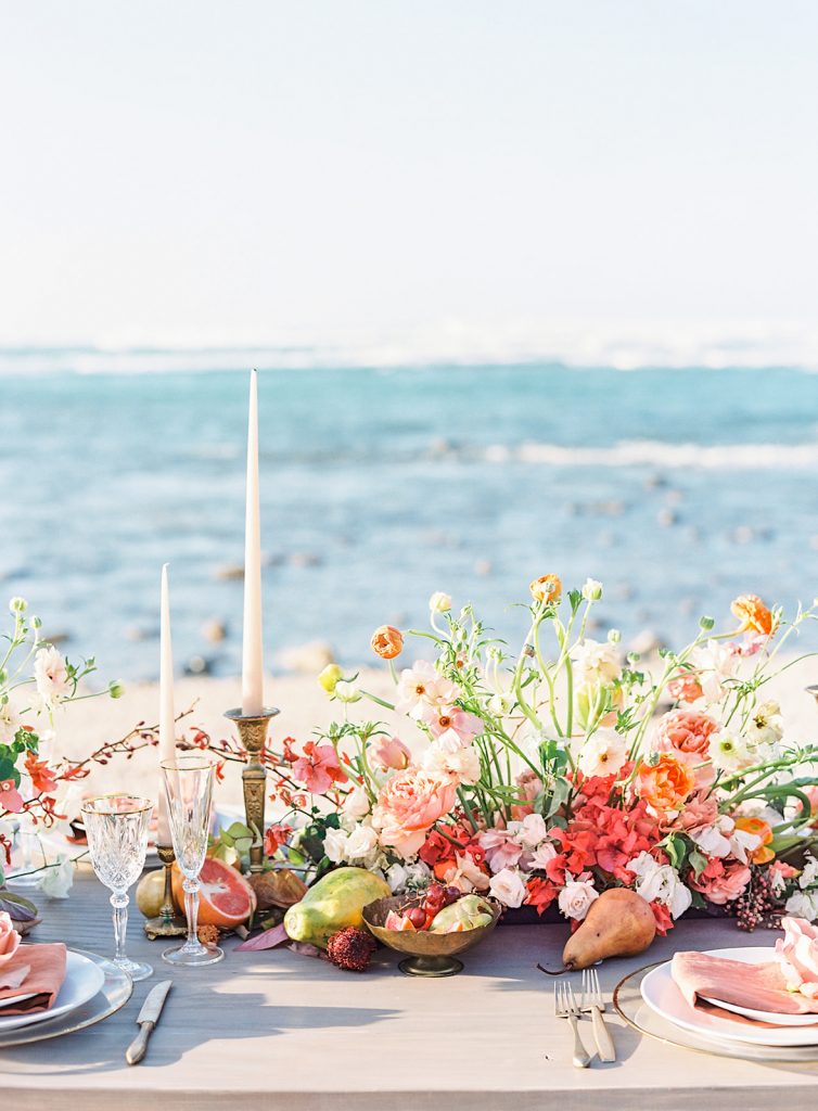 Bright vibrant orange, coral, yellow and green floral arrangements with gold candlesticks with white tapper candles set on a wedding reception table by the ocean. Ocean in the background is a rich teal and blue with some waves cresting. Photographed at destination wedding in Hawaii by Amy Mulder Photography.