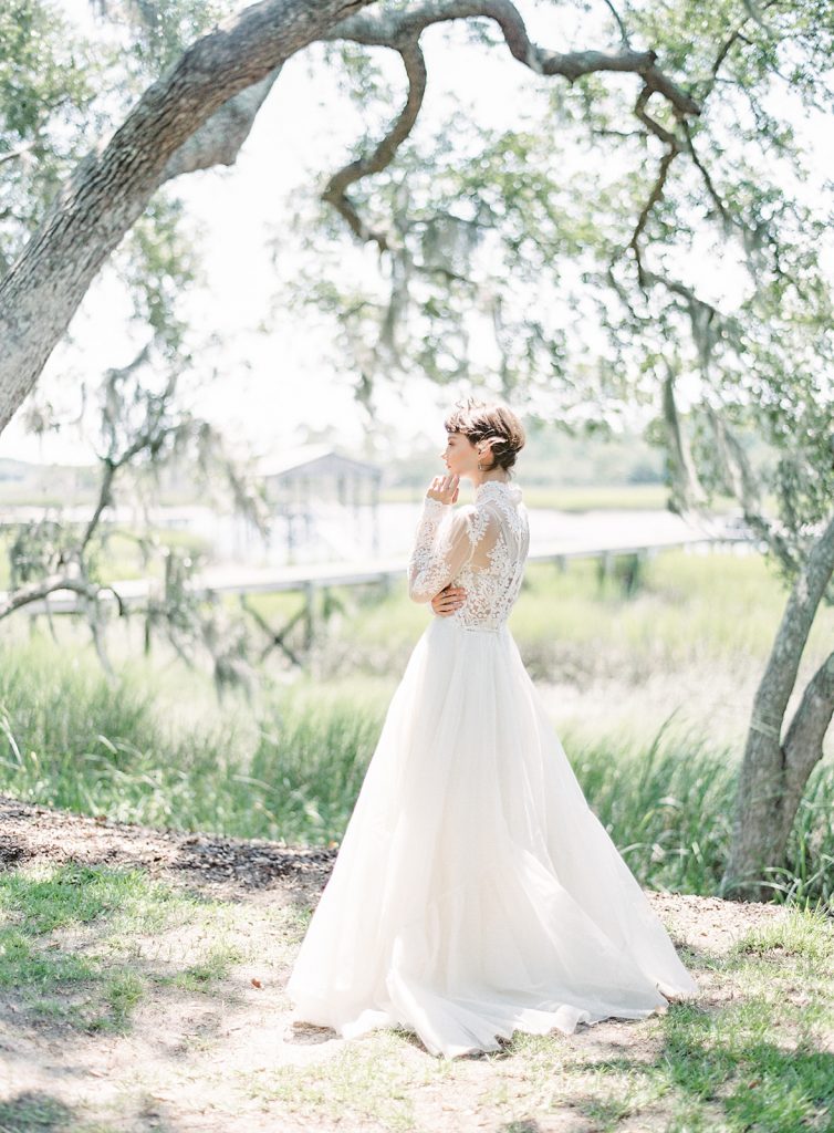 Bride standing in the sunlight at a 45degree angle from the camera. Full body photograph of bride in all lace gown under oak trees. Photographed by wedding photographers in Charleston Amy Mulder Photography.