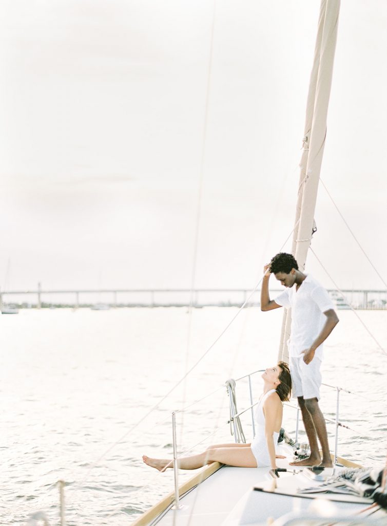 Couple on sailboat, she is sitting down with her legs over the side of the boat kicking in the wind while look up at him who is standing over her with his white shorts and shirt looking down at her. Photographed by wedding photographers in Charleston Amy Mulder Photography.