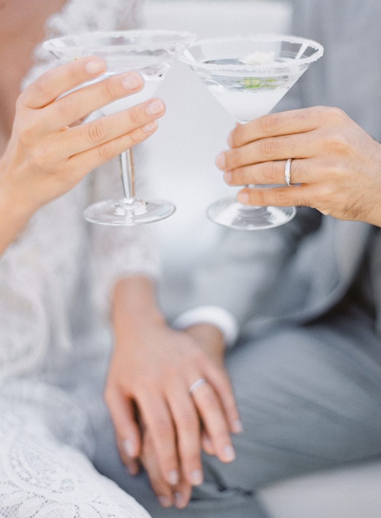 Couples hands folded on top of each other in the background with their other hands in focus at the top of the frame holding on to martini glasses with special wedding day cocktails in them. Photographed at a destination wedding in Italy by Amy Mulder Photography.