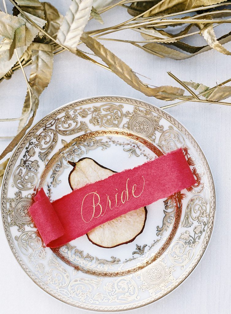 ivory plate with intricate gold detailing with dried pear slice and a red scroll with 'bride' written in gold on top. Photographed by wedding photographers in Charleston Amy Mulder Photography.