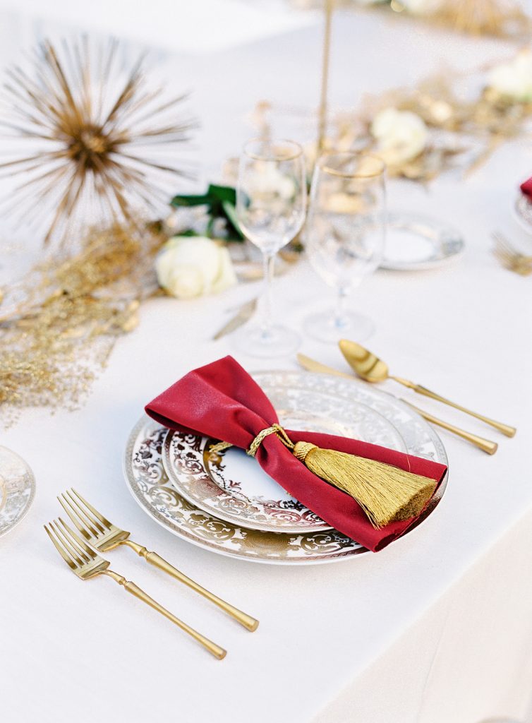 Wedding reception table with gold centerpieces of gold painted myrtle leaves and babys breath, gold candlesticks, brushed gold flatware, ivory plates with intricate gold detailing with a red velvet napkin on top with a gold tassel tied around it. Photographed at Lowndes Grove Wedding by wedding photographers in Charleston Amy Mulder Photography.
