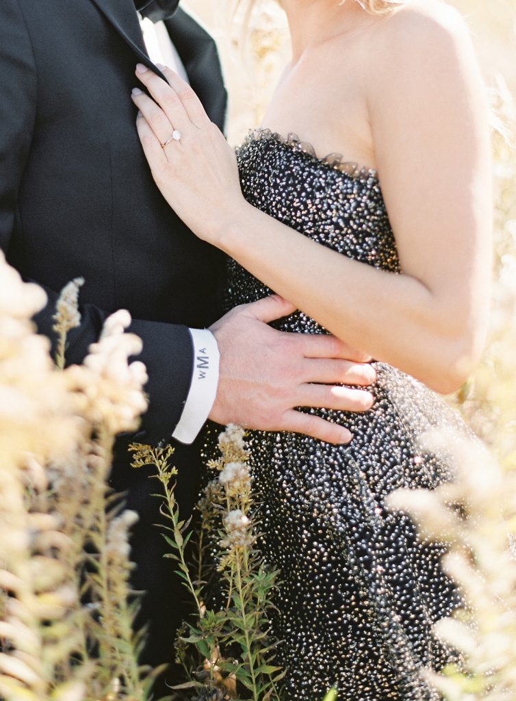 Couple standing in a tall grass field with him in a black tuxedo and monogramed tuxedo shirt cuff on her waist, her hands are on his chest. Photographed from their shoulders down to mid thigh. Photographed by wedding photographers in Charleston Amy Mulder Photography.