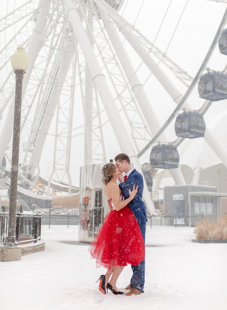 Couple in front of giant ferris wheel on Navy Pier with thick snow falling. She is in a read custom made Mira Couture gown with black Louis Vuittons, he is in a blue suit with brown shoe   Photographed in Chicago by wedding photographers in Charleston Amy Mulder Photography.