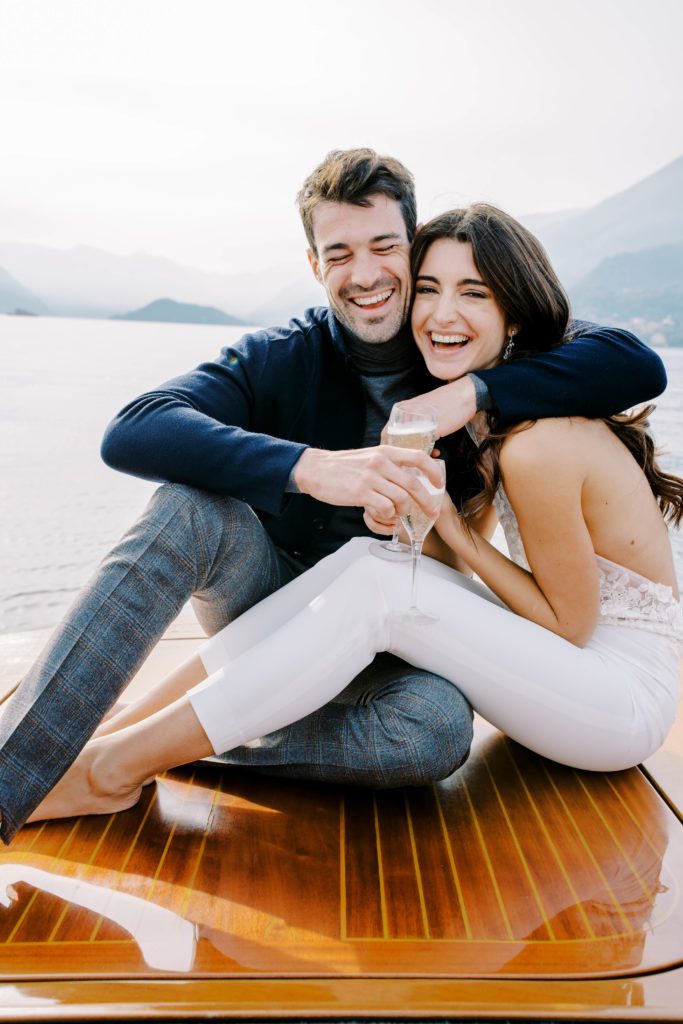Couple on back of luxury boat from Grand Hotel Tremezzo drinking prosecco and laughing. Photographed by Italy Wedding Photographer, Amy Mulder Photography.