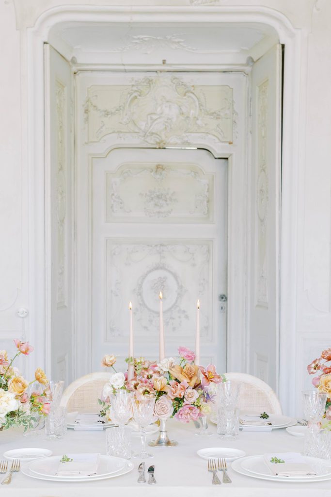 Reception table with pink, coral, purples, ivory and taupe centerpieces, white tableware, clear glass stemware with intricate stucco door behind table. Photographed by Italy wedding photographer, Amy Mulder Photography.