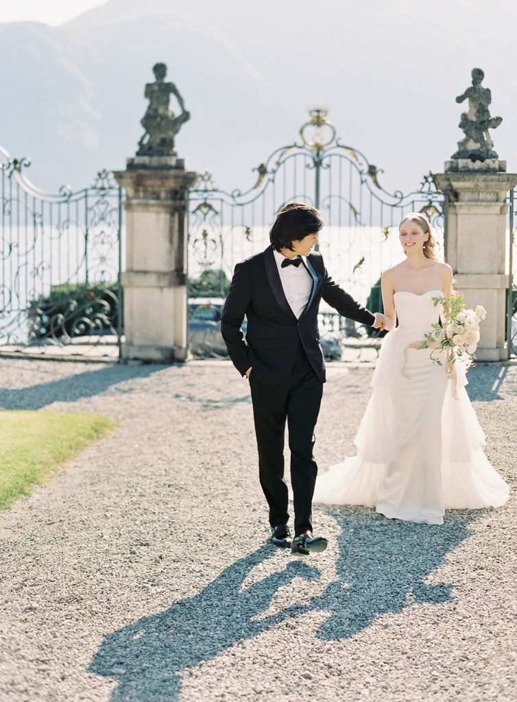 Villa Sola Cabiati Luxury wedding. Photographed by Italy wedding photographer, Amy Mulder Photography. Groom leading his bride and looking back at her as they walk towards the villa with the large main gate and Lake Como behind them.
