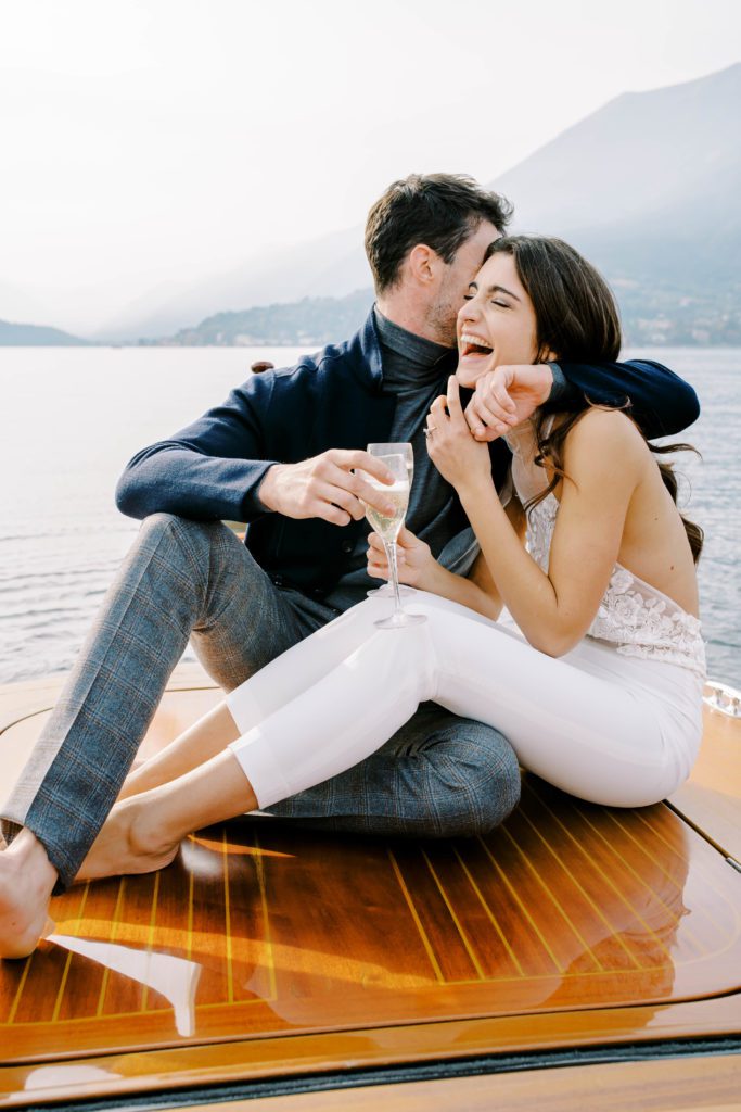 Couple on back of luxury boat from Grand Hotel Tremezzo drinking prosecco and laughing. Photographed by Italy Wedding Photographer, Amy Mulder Photography.