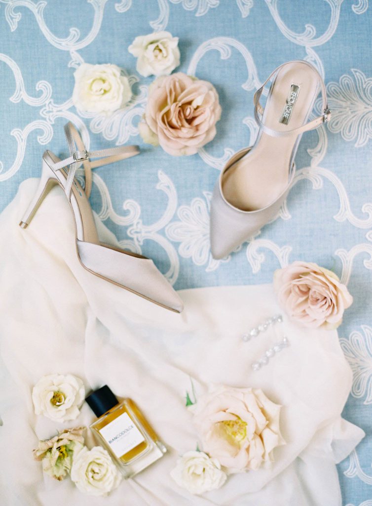 High heel shoes with perfume and florals on blue with white lace backdrop. Photographed by Italy wedding photographer, Amy Mulder Photography.
