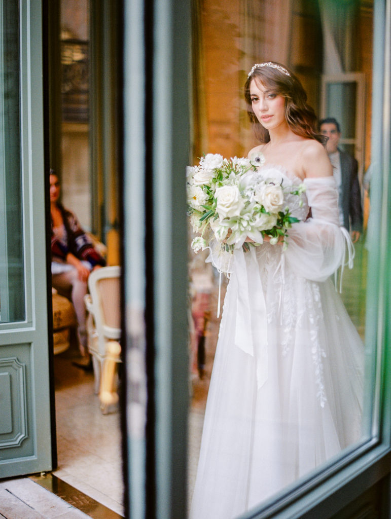Bride holding her wedding bouquet of all white flowers in a strapless lace and beaded gown with sheer long sleeves at her wedding ceremony at the Ritz Paris. 