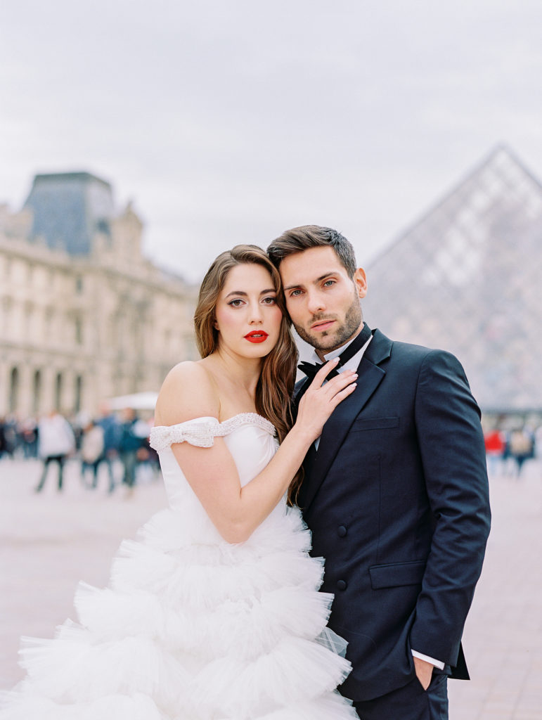 Destination wedding in Paris with bride in off the shoulder gown and groom in black tuxedo in front of the glass triangle at The Louvre.