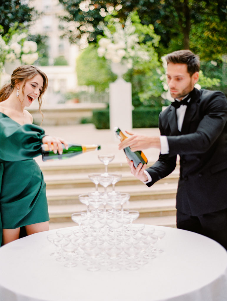 Bride in emerald green gown and groom in peach dinner jacket pouring champagne in their champagne tower for their cocktail hour at their wedding at the Ritz Paris.