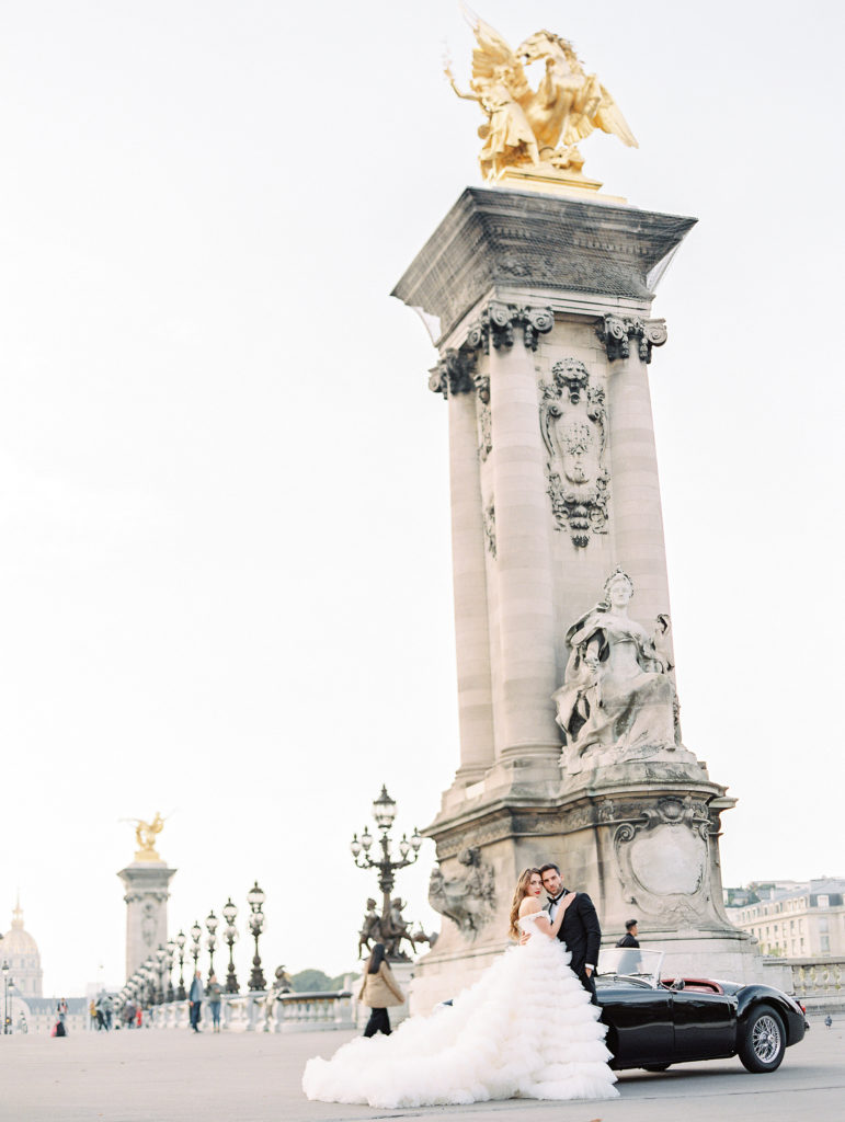 Destination wedding in Paris with bride in off the shoulder full ruffled skirt gown and groom in black tuxedo next to black vintage car on Alexander III Bridge