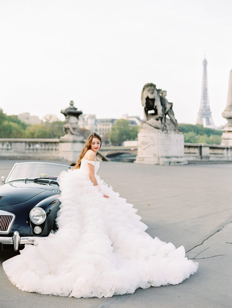 Destination wedding in Paris with bride in off the shoulder full ruffled skirt gown next to black vintage car on Alexander III Bridge with the Eiffel Tower in the background 