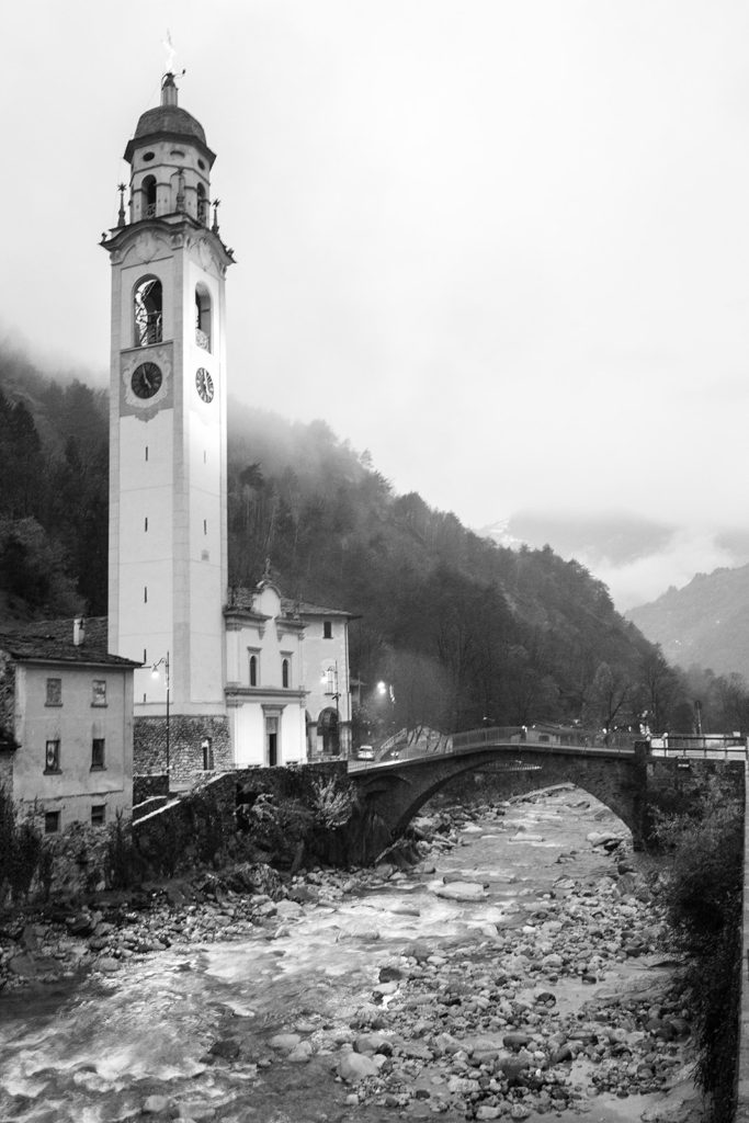 Black and white photograph of church along river in the Alps on the Italy and Swtizerland border
