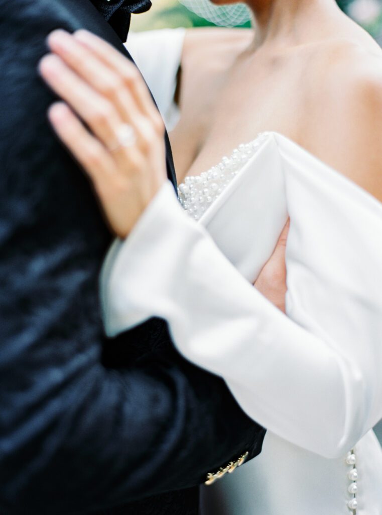 Groom in custom made textured black tuxedo embracing his wife in an off the shoulder wedding gown with buttons down the sleeves and pearls adorning the inside bust and neckline for an intimate wedding on Lake Como in Italy photographed on film by Lake Como wedding photographer