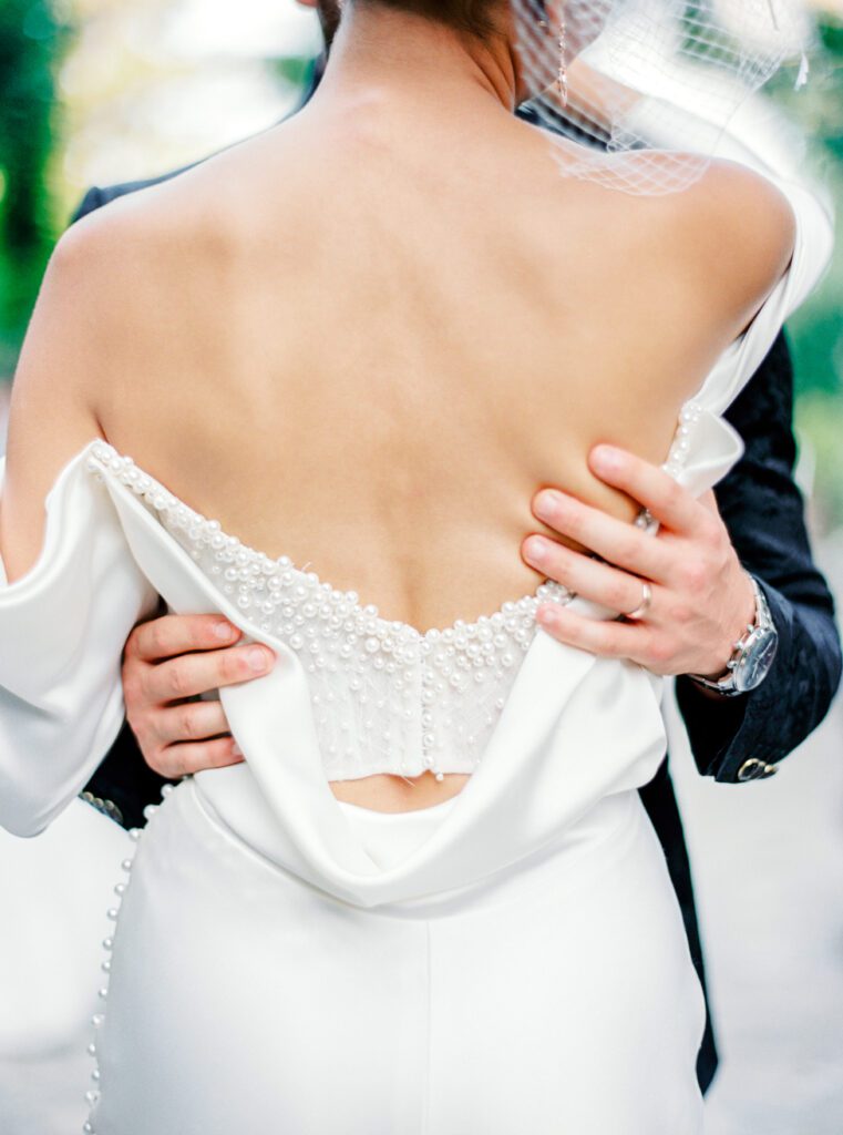 Groom in custom made textured black tuxedo embracing his wife in an off the shoulder wedding gown with buttons down the sleeves and pearls adorning the back for an intimate wedding on Lake Como in Italy photographed on film by Lake Como wedding photographer