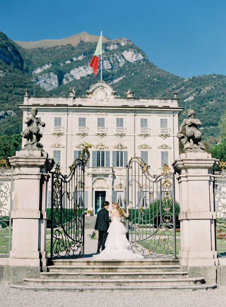 Bride and groom at the front gates of  Villa Sola Cabiati with views of the lake and mountains in the background on their wedding day at Villa Sola Cabiati on Lake Como in Italy photographed on film by Lake Como wedding photographer