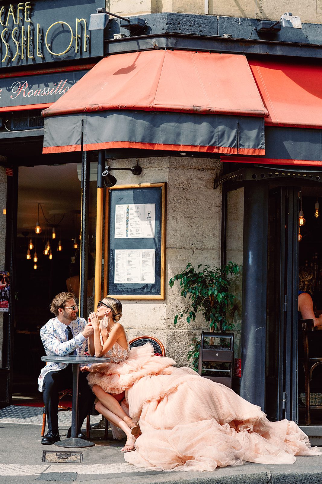 A Paris Engagement Session couple in formal attire, he in black dress trousers and floral patterned dinner jacket and she in a hi-low peach deep v neck and multi tiered gown with the couple laughing and holding hands while sitting at a bistro table together outside a cafe photographed by Italy wedding photographer.