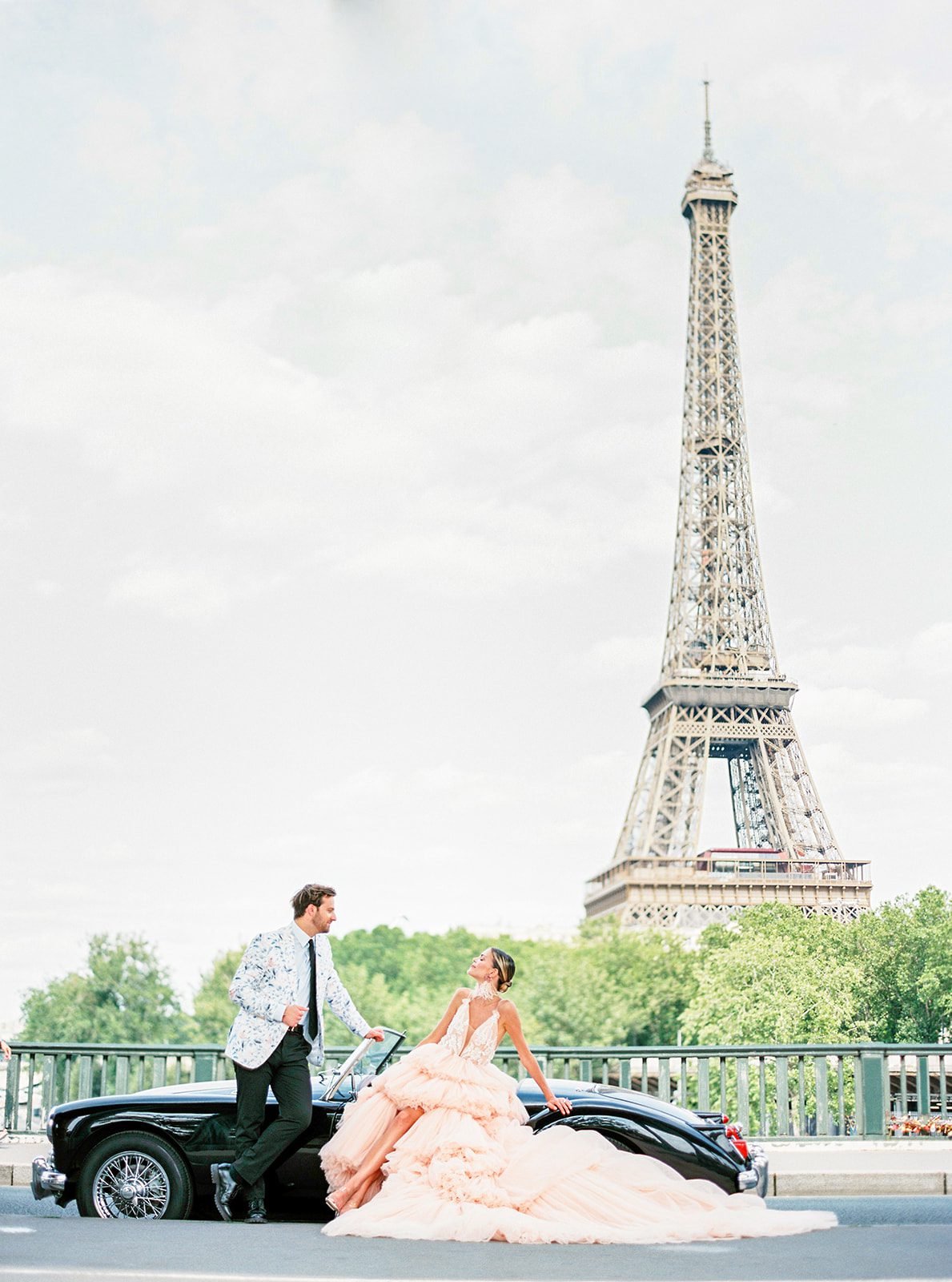 A Paris Engagement Session with vintage black convertible, couple in formal attire, he in black dress trousers and floral patterned dinner jacket and she in a hi-low peach deep v neck and multi tiered gown standing with the Eiffel Tower in the background photographed on film in Paris by Italy Wedding Photographer