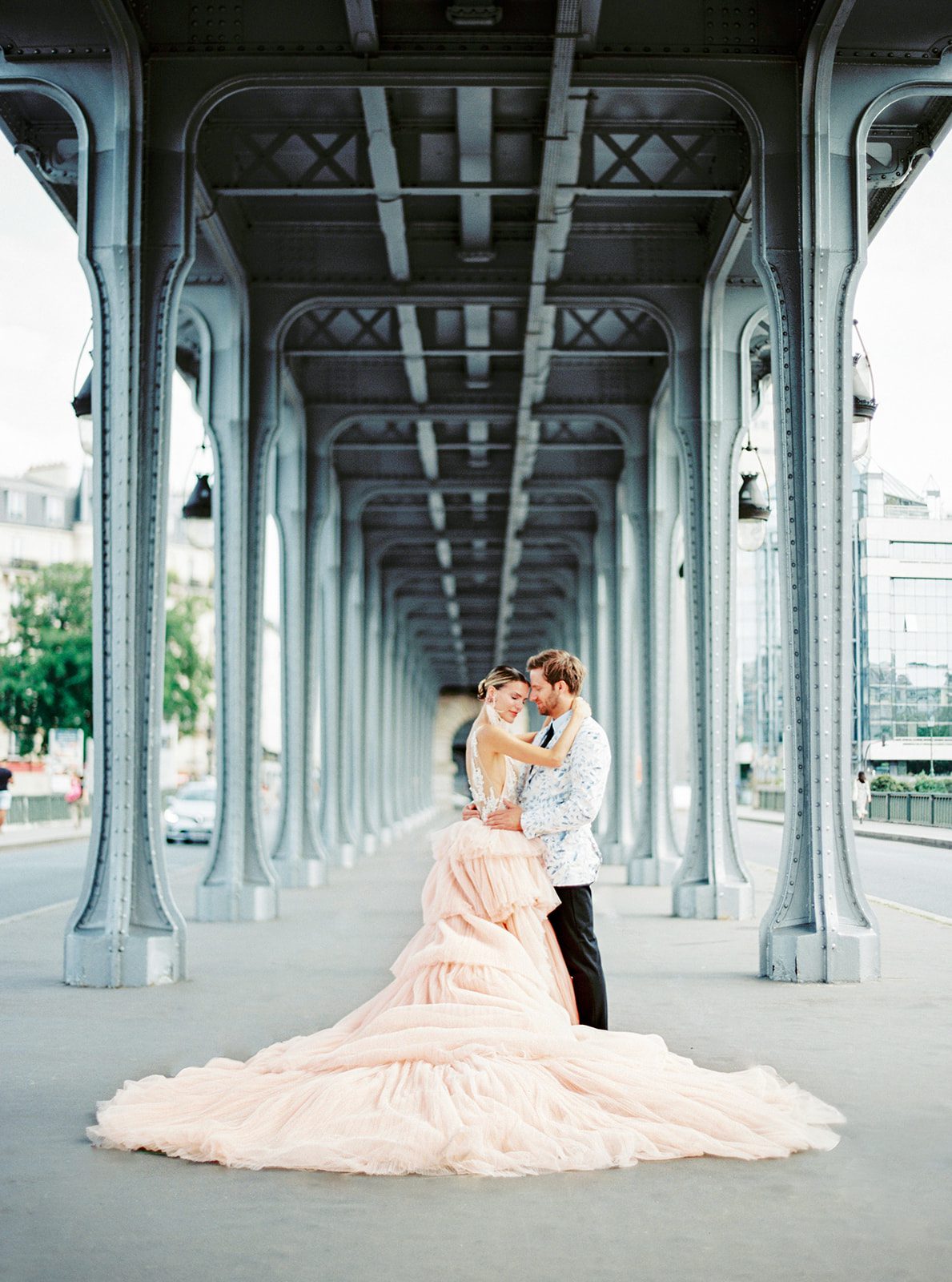 A Paris Engagement Session couple in formal attire, he in black dress trousers and floral patterned dinner jacket and she in a hi-low peach deep v neck and multi tiered gown standing foreheads together and arms wrapped around each other with grey/blue archways leading down the walkway photographed on film in Paris by Italy Wedding Photographer