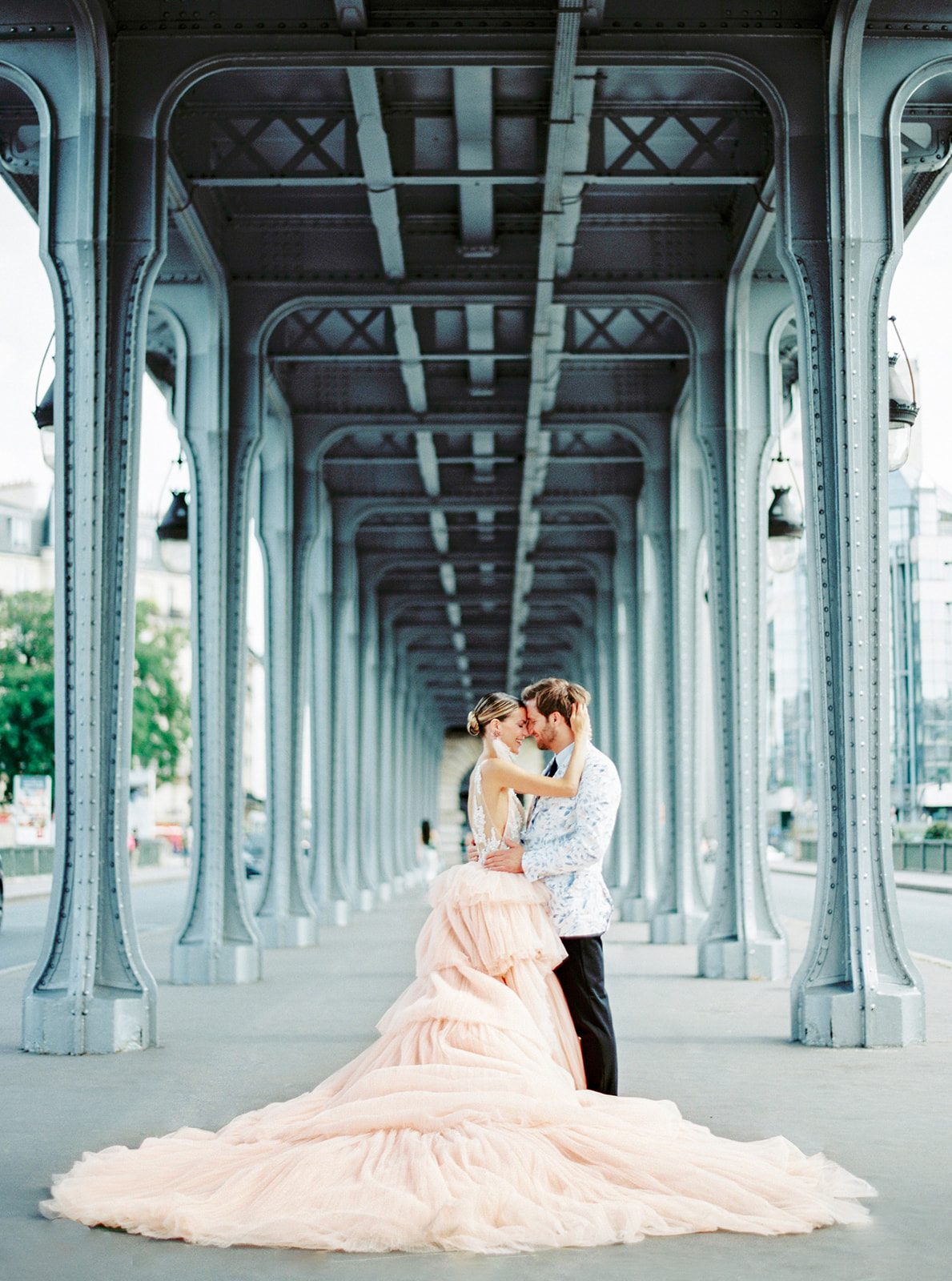 A Paris Engagement Session couple in formal attire, he in black dress trousers and floral patterned dinner jacket and she in a hi-low peach deep v neck and multi tiered gown standing foreheads together and arms wrapped around each other with grey/blue archways leading down the walkway photographed on film in Paris by Italy Wedding Photographer