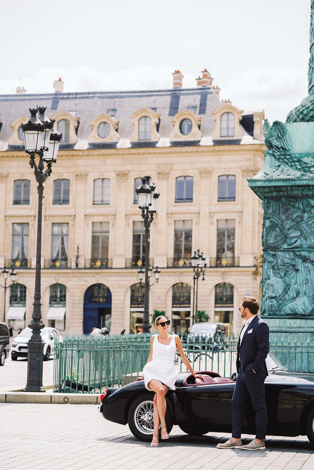 Paris engagement session at Place Vendome outside of the Ritz Paris bride is in a short 3d patterned sleeveless dress sitting on a vintage black car looking at her groom who is in a navy suit photographed on film by Italy wedding photographer