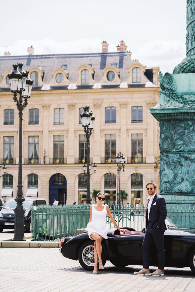 Paris engagement session at Place Vendome outside of the Ritz Paris bride is in a short 3d patterned sleeveless dress sitting on a vintage black car, her groom is in a navy suit photographed on film by Italy wedding photographer