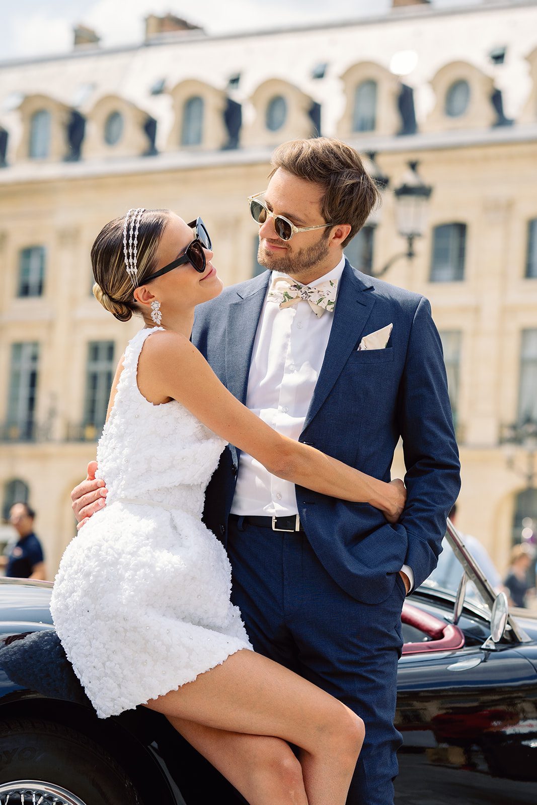 Paris engagement session at Place Vendome outside of the Ritz Paris bride is in a short 3d patterned sleeveless dress sitting on a vintage black car smiling and embracing her groom who is in a navy suit photographed on film by Italy wedding photographer