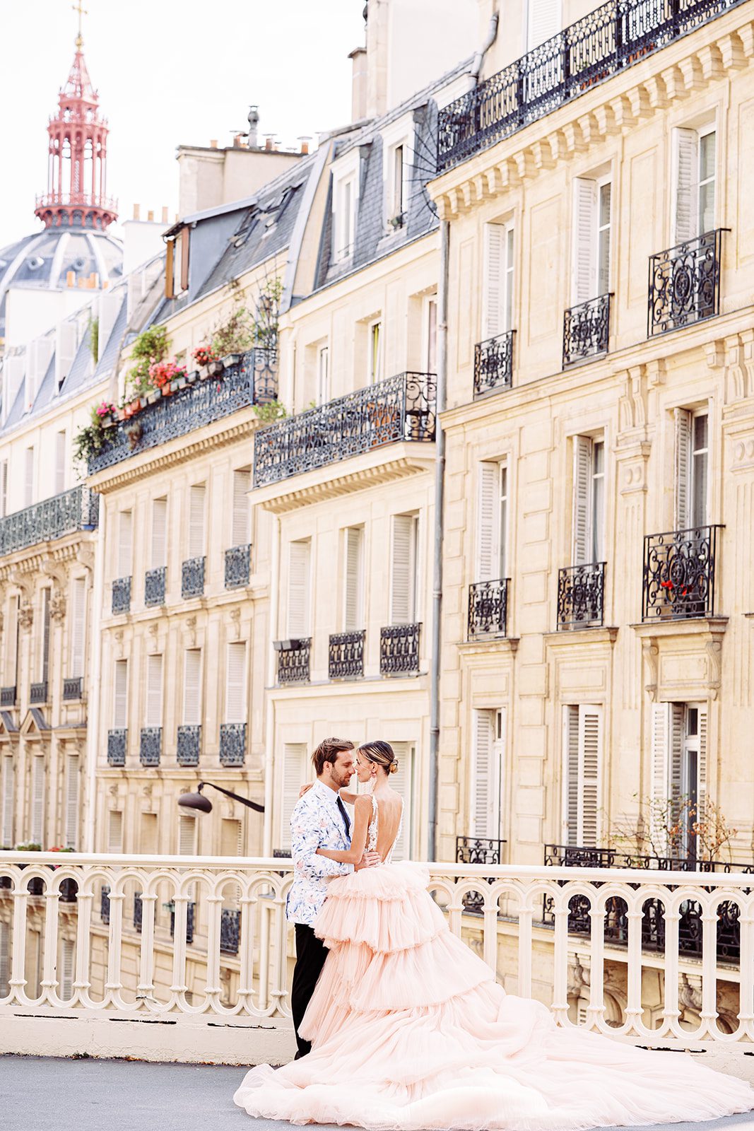 A Paris Engagement Session with couple in formal attire, he in black dress trousers and floral patterned dinner jacket and she in a hi-low peach deep v neck and multi tiered gown standing with apartments of Paris in the background photographed on film in Paris by Italy Wedding Photographer