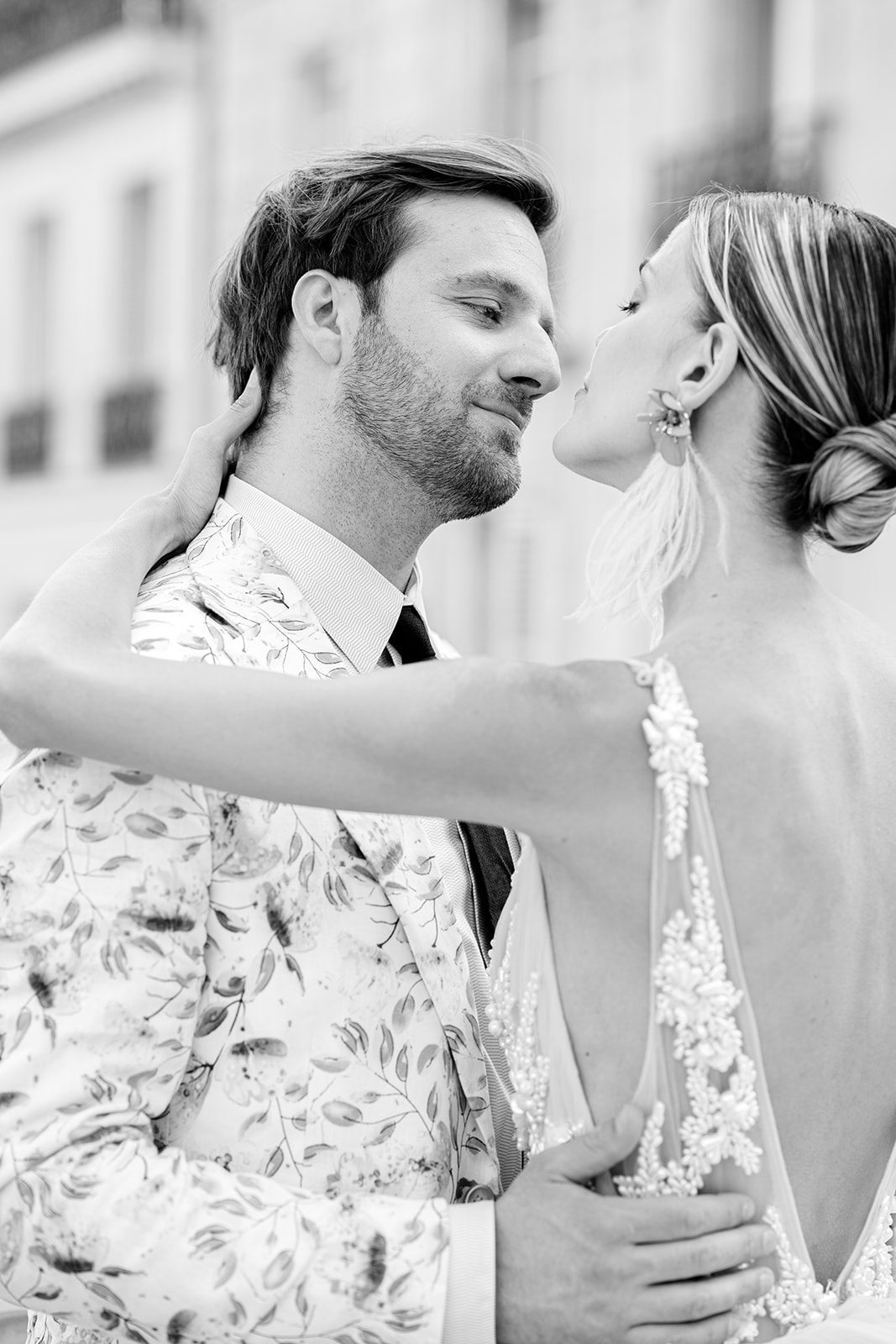 Black and white film photograph with couple in formal attire, he in black dress trousers and floral patterned dinner jacket and she in a hi-low peach deep v neck and multi tiered gown standing with apartments of Paris in the background photographed on film in Paris by Italy Wedding Photographer