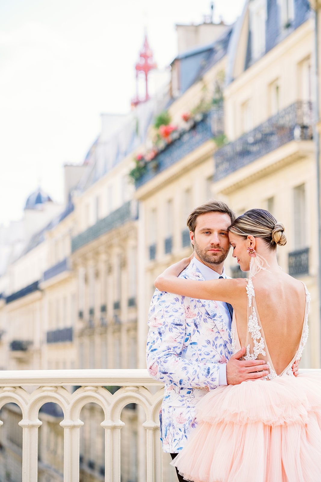 A Paris Engagement Session with couple in formal attire, he in black dress trousers and floral patterned dinner jacket and she in a hi-low peach deep v neck and multi tiered gown standing with apartments of Paris in the background photographed on film in Paris by Italy Wedding Photographer