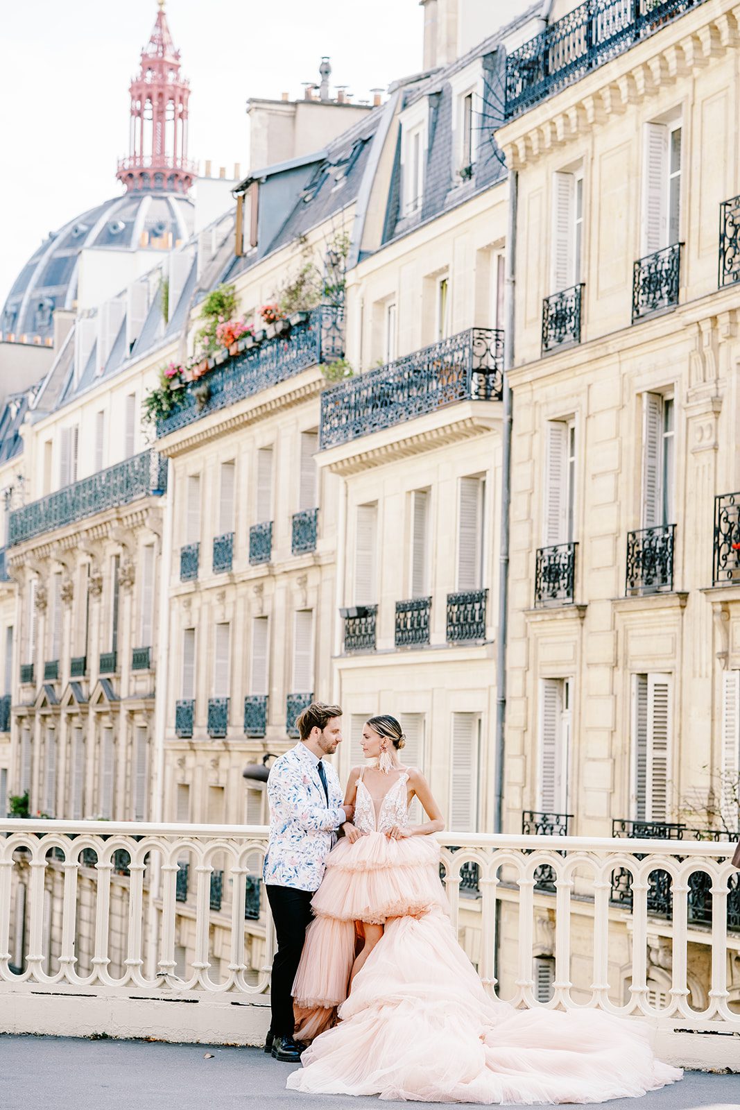 A Paris Engagement Session with couple in formal attire, he in black dress trousers and floral patterned dinner jacket and she in a hi-low peach deep v neck and multi tiered gown standing with apartments of Paris in the background photographed on film in Paris by Italy Wedding Photographer