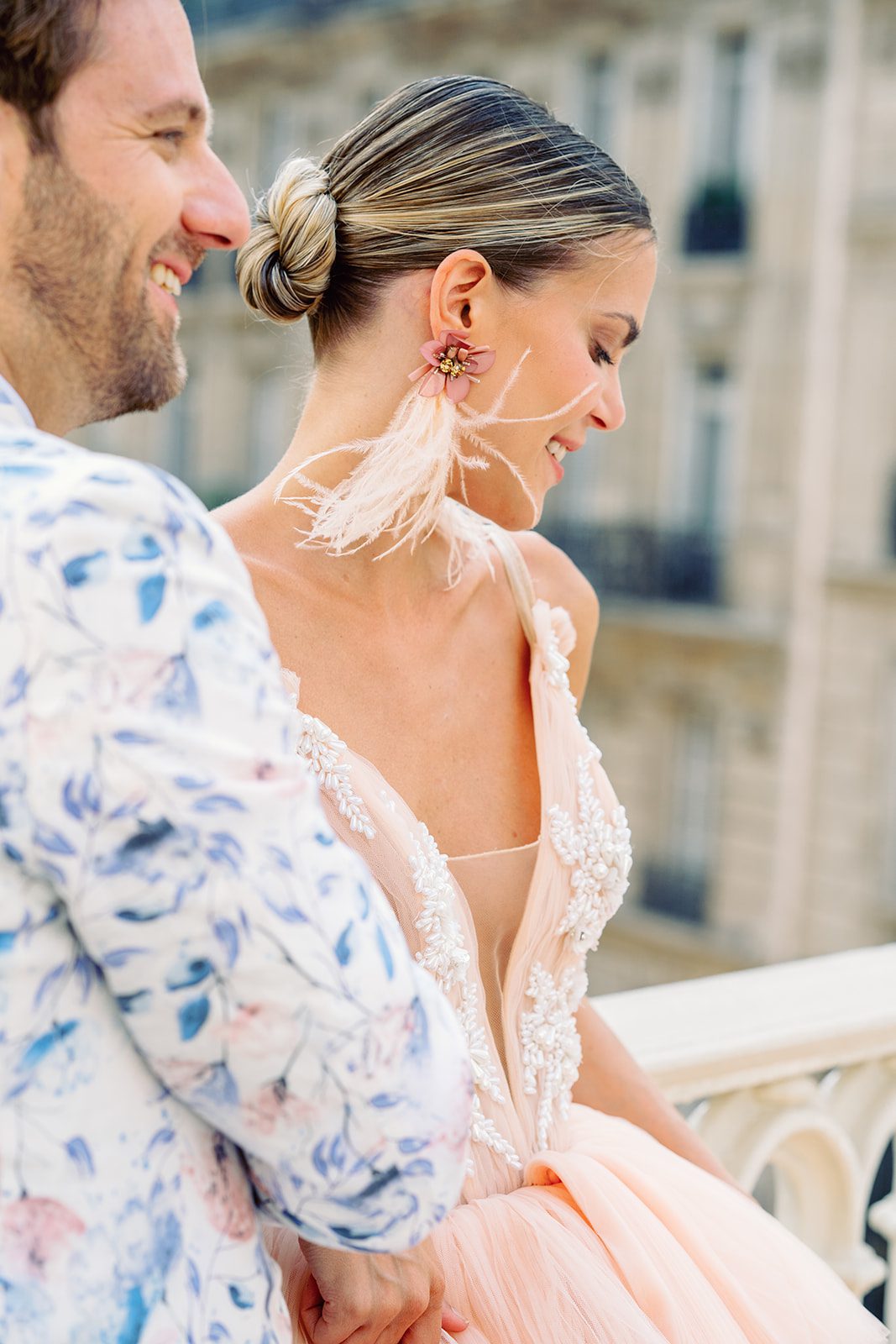 A Paris Engagement Session with couple in formal attire, he in black dress trousers and floral patterned dinner jacket and she in a hi-low peach deep v neck and multi tiered gown standing with apartments of Paris in the background photographed in Paris by Italy Wedding Photographer