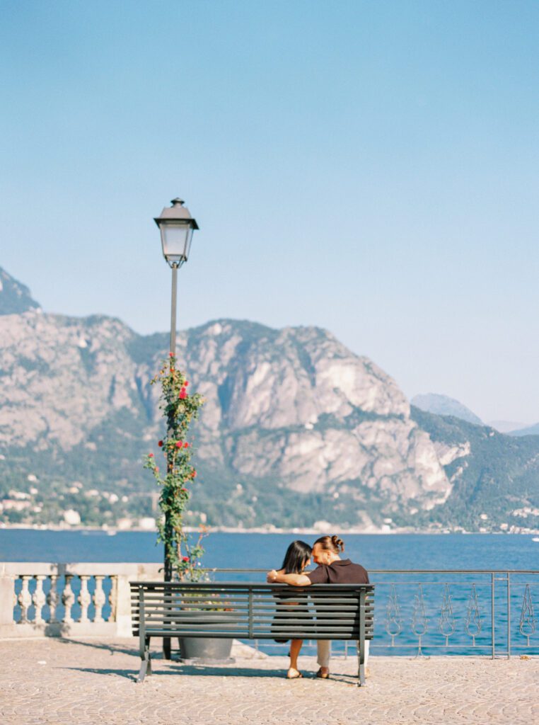 Couple sitting on bench by Lake Como, showing the bright blue of the lake and mountains in the back photographed by Lake Como Photographer