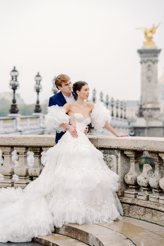 Paris pre-wedding session on Pont Alexandre III with bride in full train layered gown with separate off the shoulder full puffed sleeves and groom in a navy blue suit photographed by Italy destination photographer