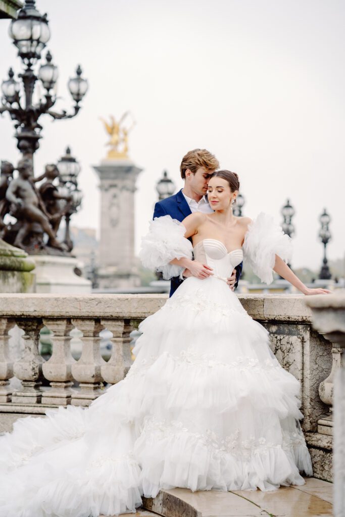 Paris pre-wedding session on Pont Alexandre III with bride in full train layered gown with separate off the shoulder full puffed sleeves and groom in a navy blue suit photographed by Italy destination photographer