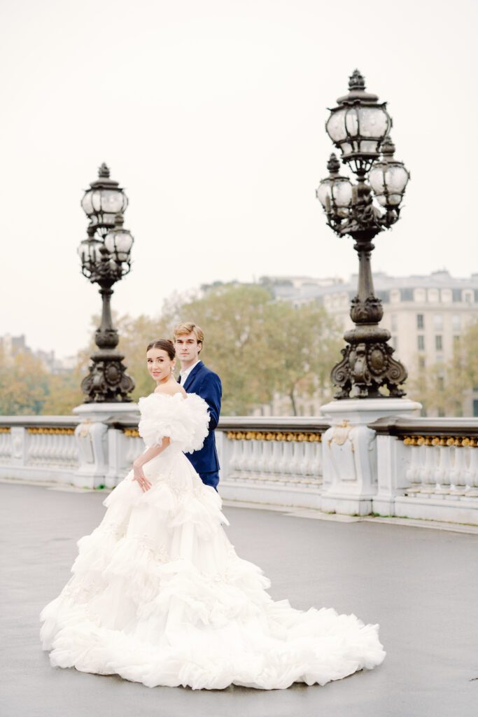 Paris pre-wedding session on Pont Alexandre III with bride in full train layered gown with separate off the shoulder full puffed sleeves and groom in a navy blue suit photographed by Italy destination photographer