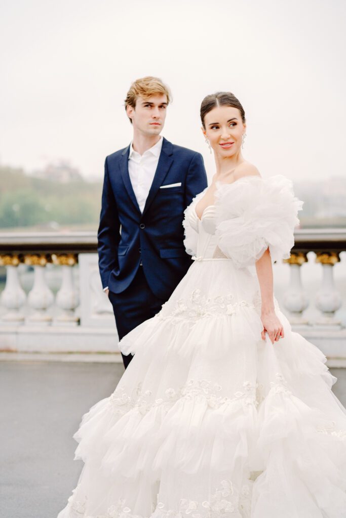 Paris pre-wedding session on Pont Alexandre III with bride in full train layered gown with separate off the shoulder full puffed sleeves and groom in a navy blue suit photographed by Italy destination photographer